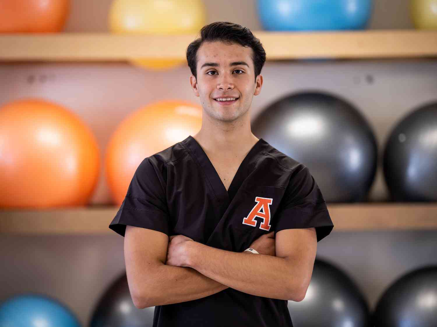 Estudiante con uniforme negro y logotipo de la A posa sonriente con brazos cruzados frente a pelotas de ejercicio en un aula de rehabilitación o fisioterapia.